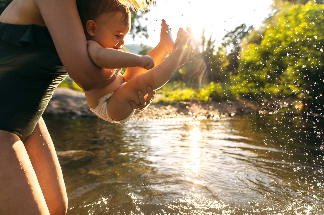 An adult holds a baby over a sunlit river, splashing water. The scene is outdoors with greenery in the background.