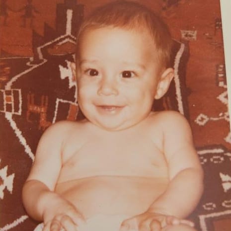 A smiling baby with short hair sits on a patterned carpet, looking slightly off to the side. The photo has a sepia tone.