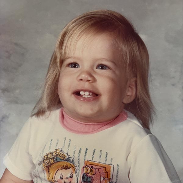 Toddler with light hair and blue eyes smiles, wearing a white shirt with cartoon characters, posing for a studio portrait against a gray background.