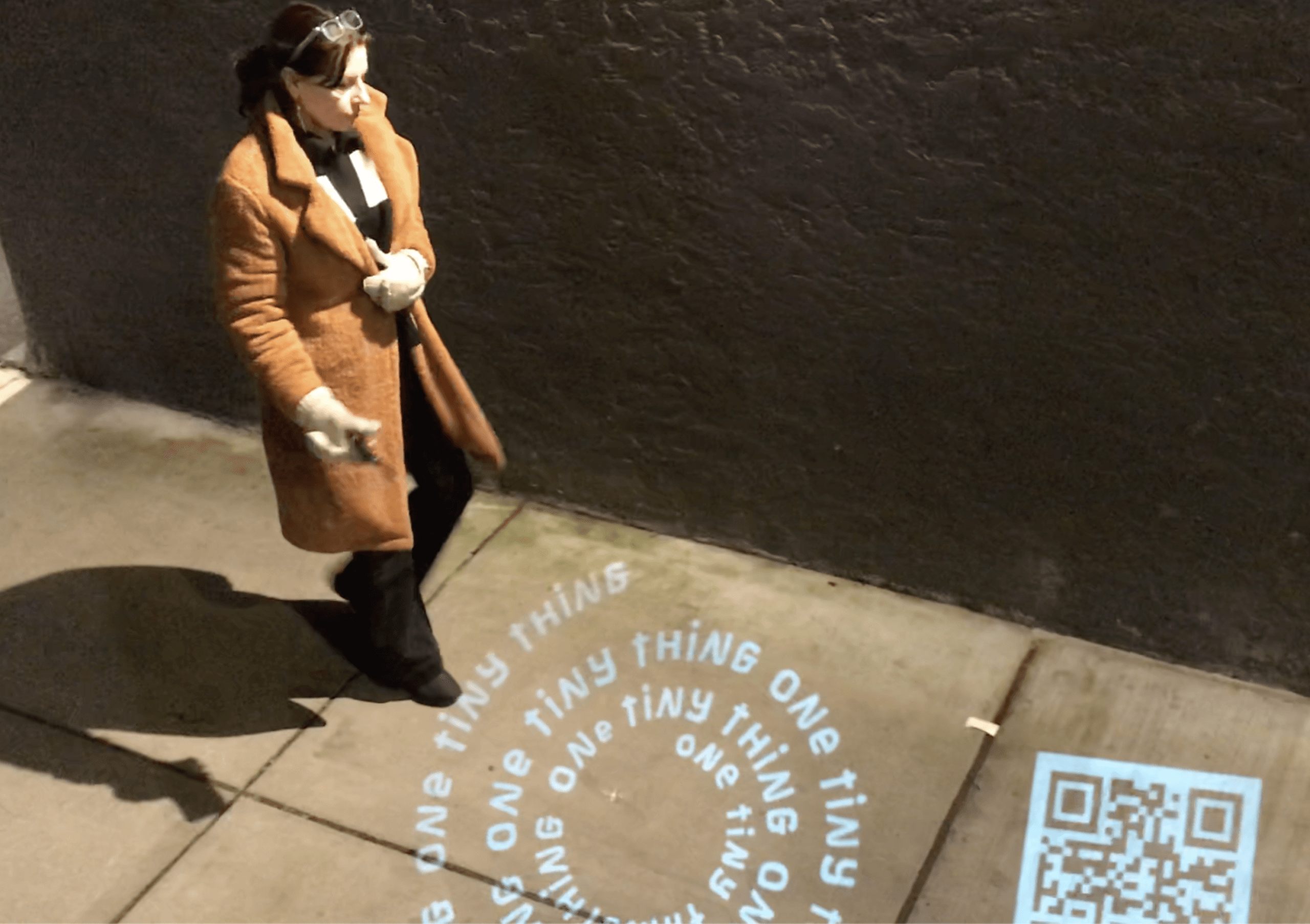 A person walks on a sidewalk at night past a projected circular text on the ground that repeats “ONE TINY THING” next to a QR code.