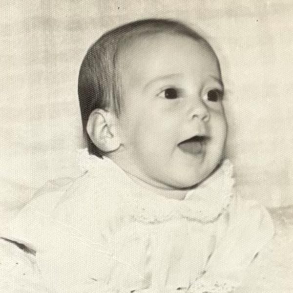 Black and white photo of a baby wearing a light-colored outfit, looking slightly upward with mouth open and eyes wide.