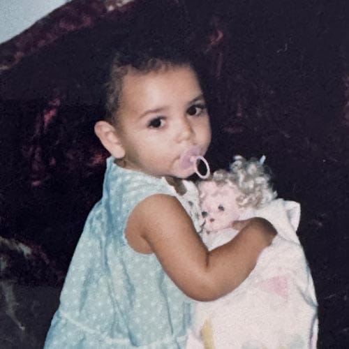 A young child in a light blue dress holds a doll and has a pacifier in their mouth, looking toward the camera.
