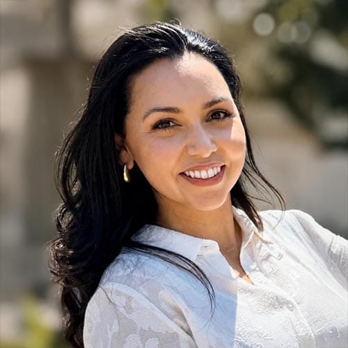 A woman with long dark hair, wearing a white blouse and gold hoop earrings, smiles outdoors in natural light.