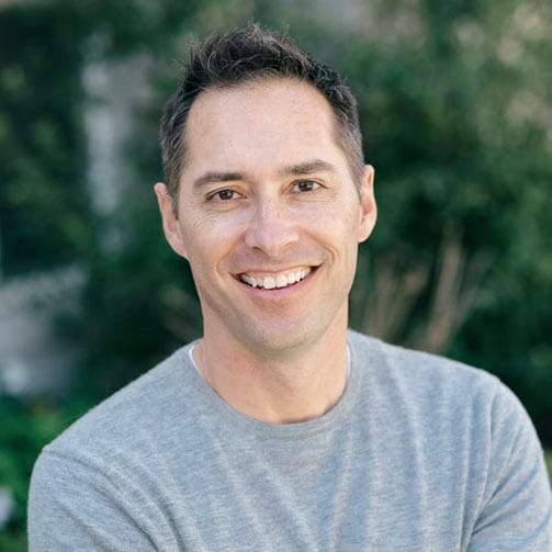 A man with short brown hair, wearing a light gray sweater, smiles at the camera with greenery blurred in the background.