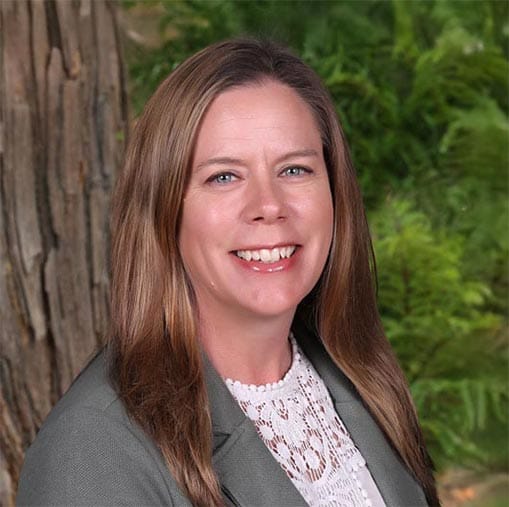 A woman with straight light brown hair, wearing a gray blazer and white lace top, smiles in front of a background of green foliage and tree bark.