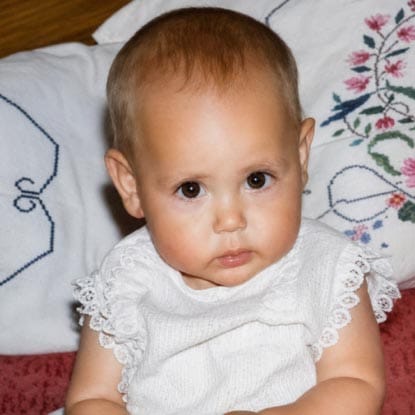 Baby with short hair and round cheeks wearing a white outfit sits in front of embroidered pillows, looking directly at the camera.
