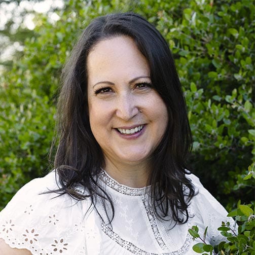 A woman with long dark hair wearing a white blouse stands outdoors in front of green bushes, smiling at the camera.