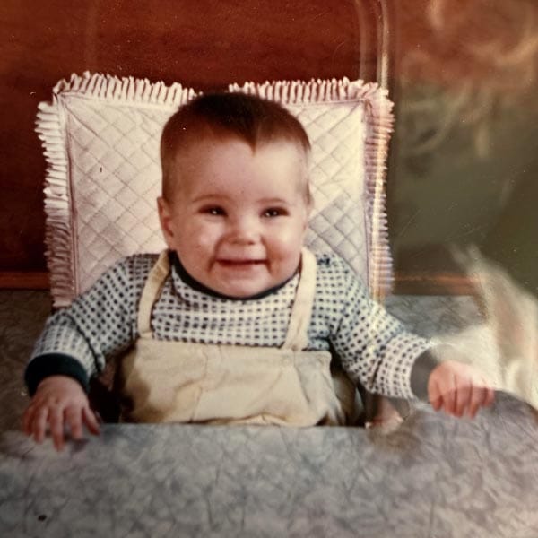 A smiling baby sits at a table, wearing a patterned long-sleeve shirt and light-colored overalls, with a quilted cushion behind them.