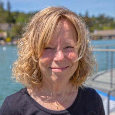 A woman with shoulder-length blonde hair stands outdoors near a body of water, smiling, with trees and houses in the background.