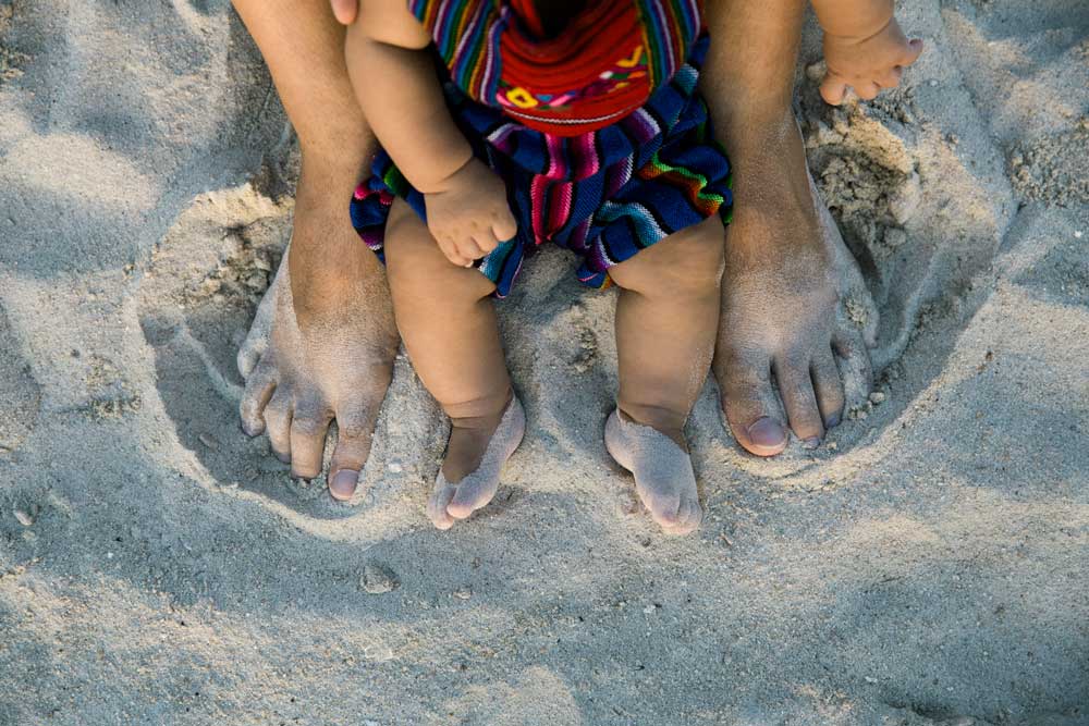 Close-up of an adult and a baby sitting on sand with their bare feet next to each other; both have sand on their legs and feet.