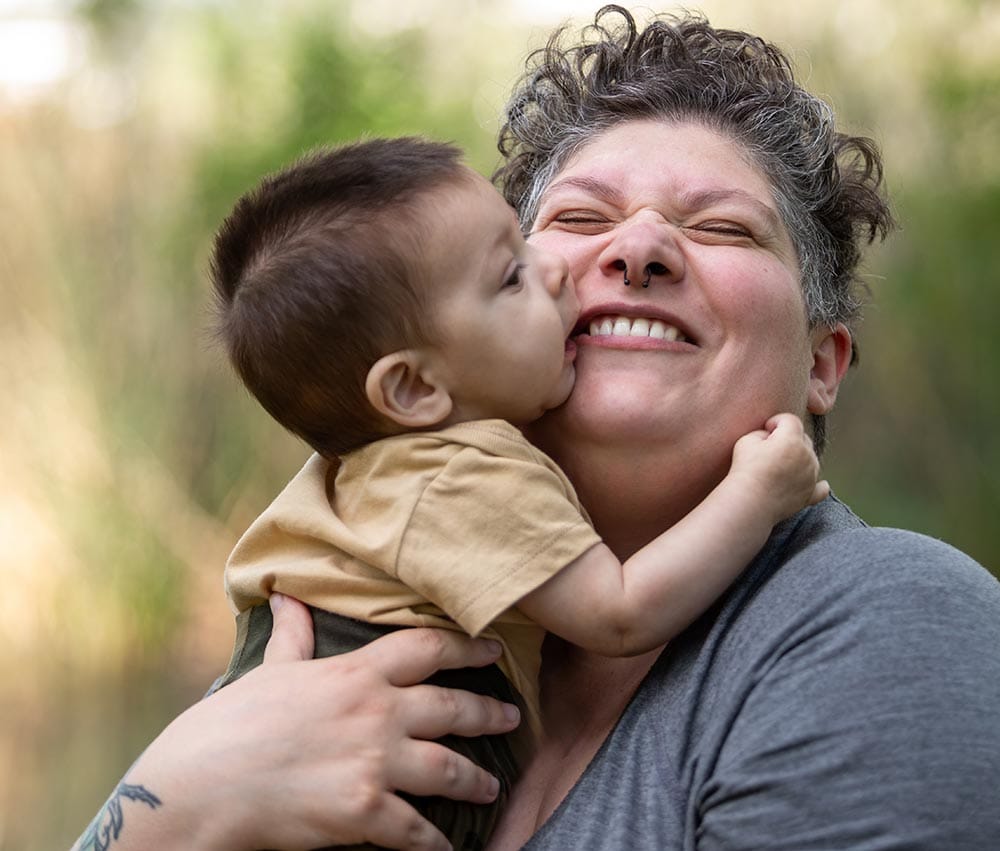 An adult holding a young child who is kissing their cheek outdoors, both appearing happy and affectionate.