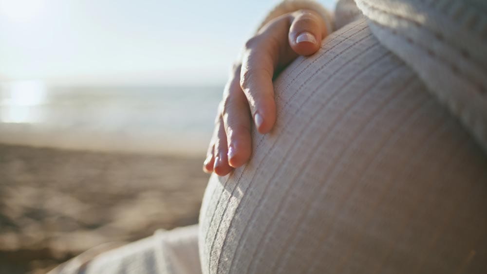 A close-up of a person's hand resting on their pregnant belly, with a blurred beach and ocean in the background.